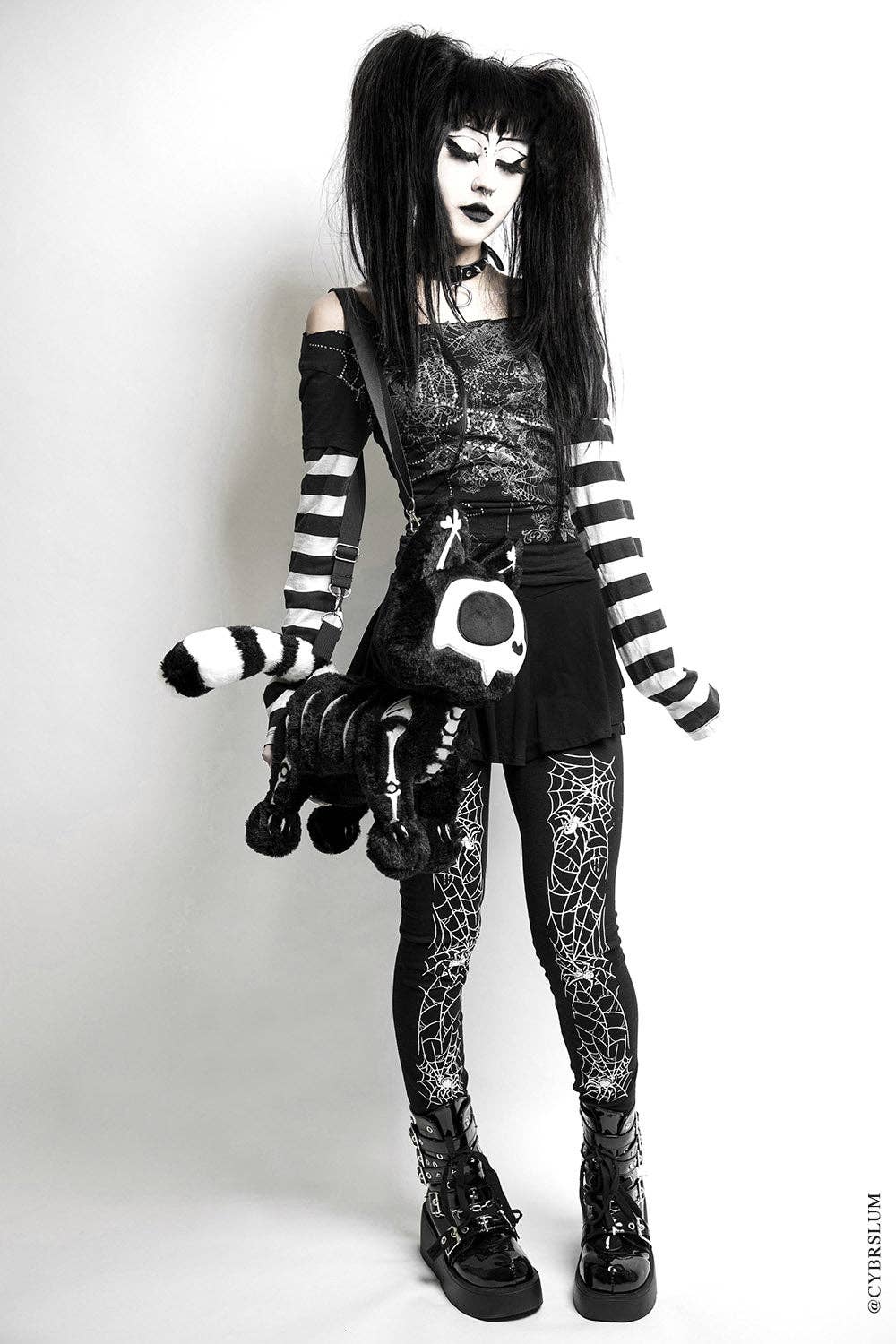 A goth-style model with long black hair and dramatic white face makeup wears a black-and-white striped top and matching striped arm warmers, a black mini skirt, spider-print tights, and black lace-up boots. She carries a black-and-white striped plush bag with a skull patch and a cat tail accessory, photographed against a plain light gray studio background.