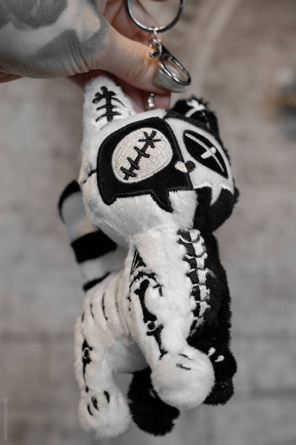A close-up of a hand holding a plush cat keychain with a metal key ring. The plush is black and white with embroidered stitched details, including a football pattern on one eye and a cross on the other.
