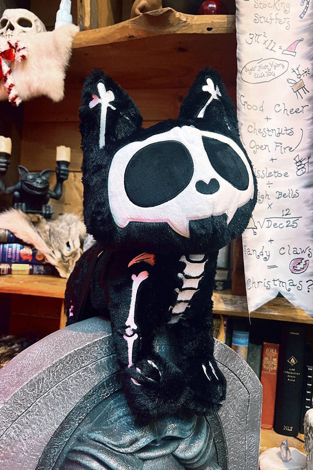 A black plush cat bag styled as a skeleton, sitting on a gray textured surface in a cluttered, cozy room with wooden shelves, books, and Halloween decorations in the background.