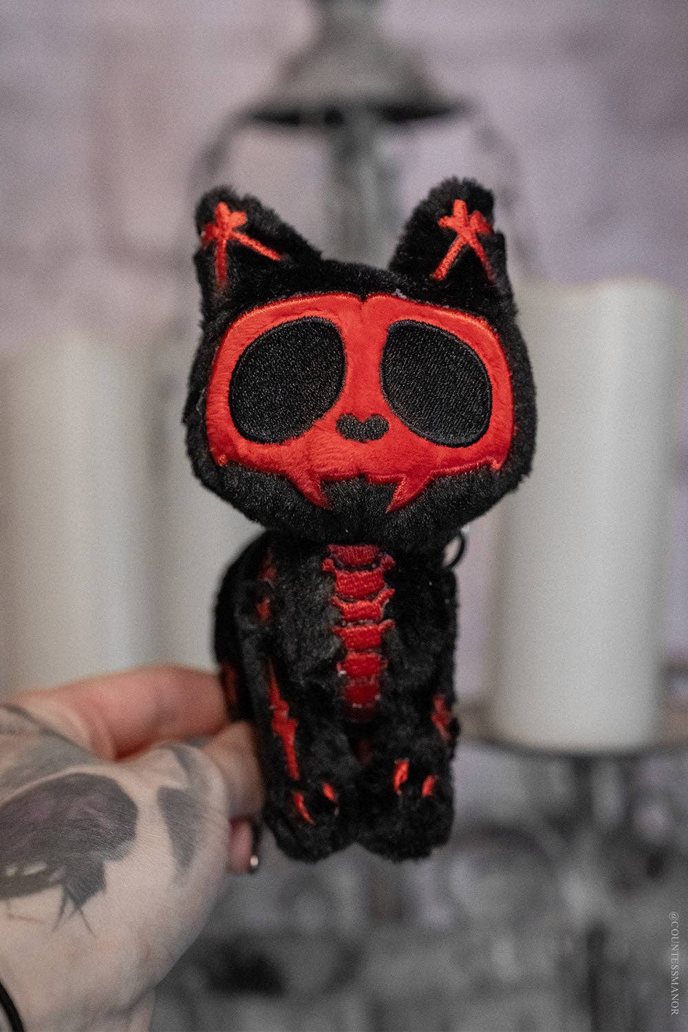 Black and red plush cat keychain held by a person with tattoos against a blurred background