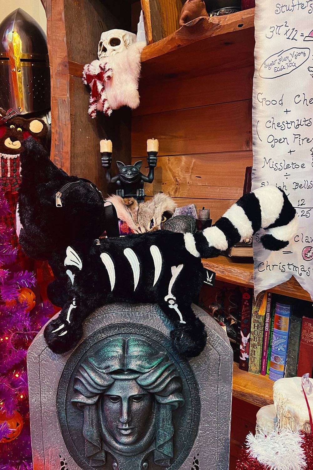 A black and white striped plush bag with white skeleton bones on its body, posed on top of a gray stone tombstone in a rustic, decorated room with shelves, books, and candles.