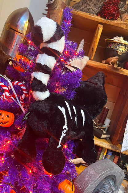 A black cat plush crossbody with white skeleton print and a striped tail is hanging on a purple tree decorated with red lights, surrounded by Halloween decorations.