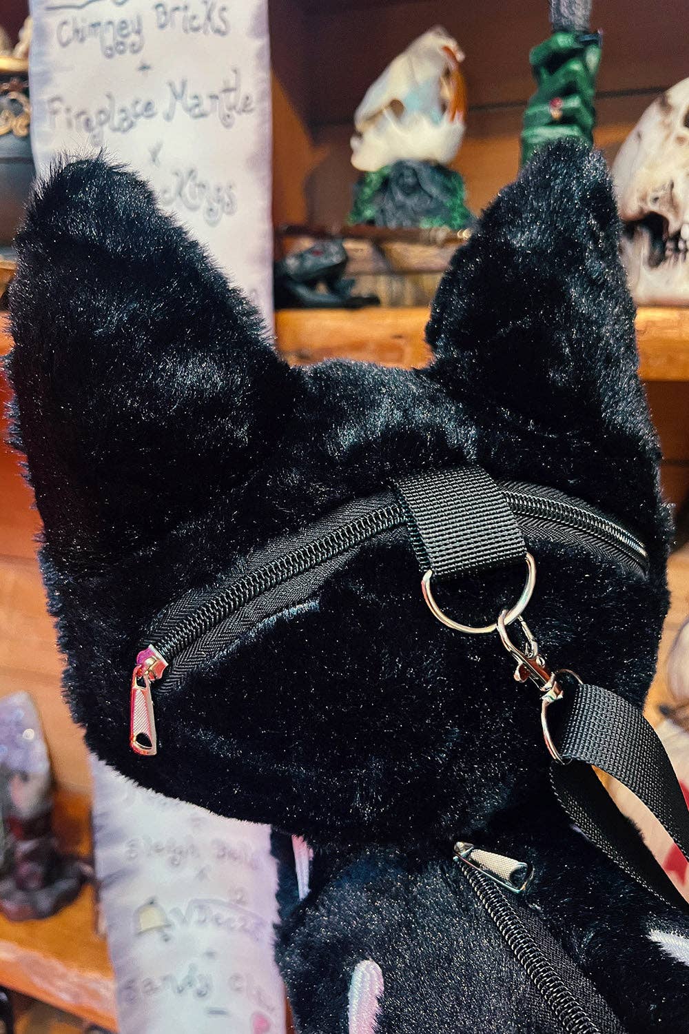 A close-up of a black plush cat bag featuring a black nylon strap with a silver metal ring and crossbody attachment, set against a blurred background of a shop interior.