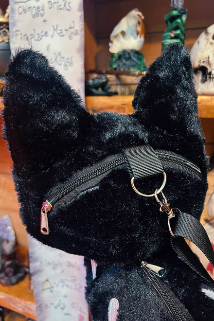 A close-up of a black plush cat bag featuring a black nylon strap with a silver metal ring and crossbody attachment, set against a blurred background of a shop interior.