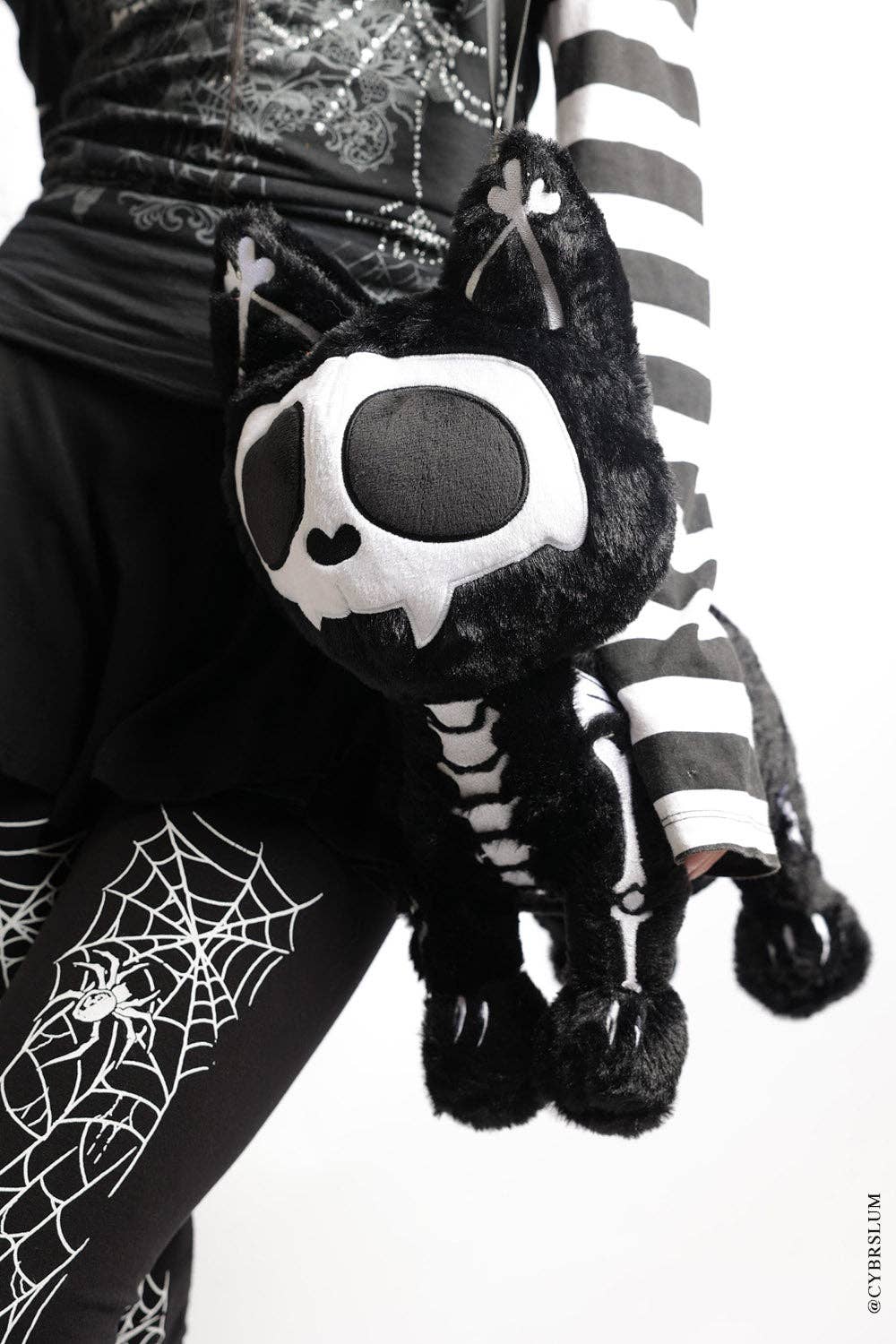 A black and white skeleton-themed plush cat bag with a skull face and tattered tail, carried on a person wearing a black and white striped top and spider-print leggings.