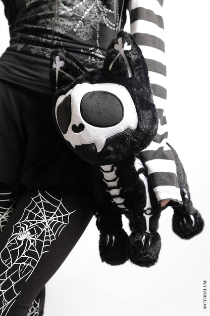 A black and white skeleton-themed plush cat bag with a skull face and tattered tail, carried on a person wearing a black and white striped top and spider-print leggings.