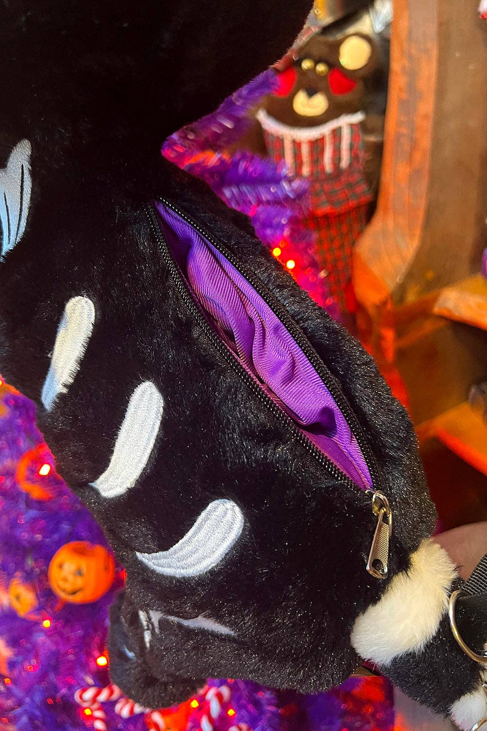 A close-up of a black plush crossbody with white embroidered details and a purple interior lining, shown with a zipper opening and a small gold zipper pull in a decorated indoor setting.