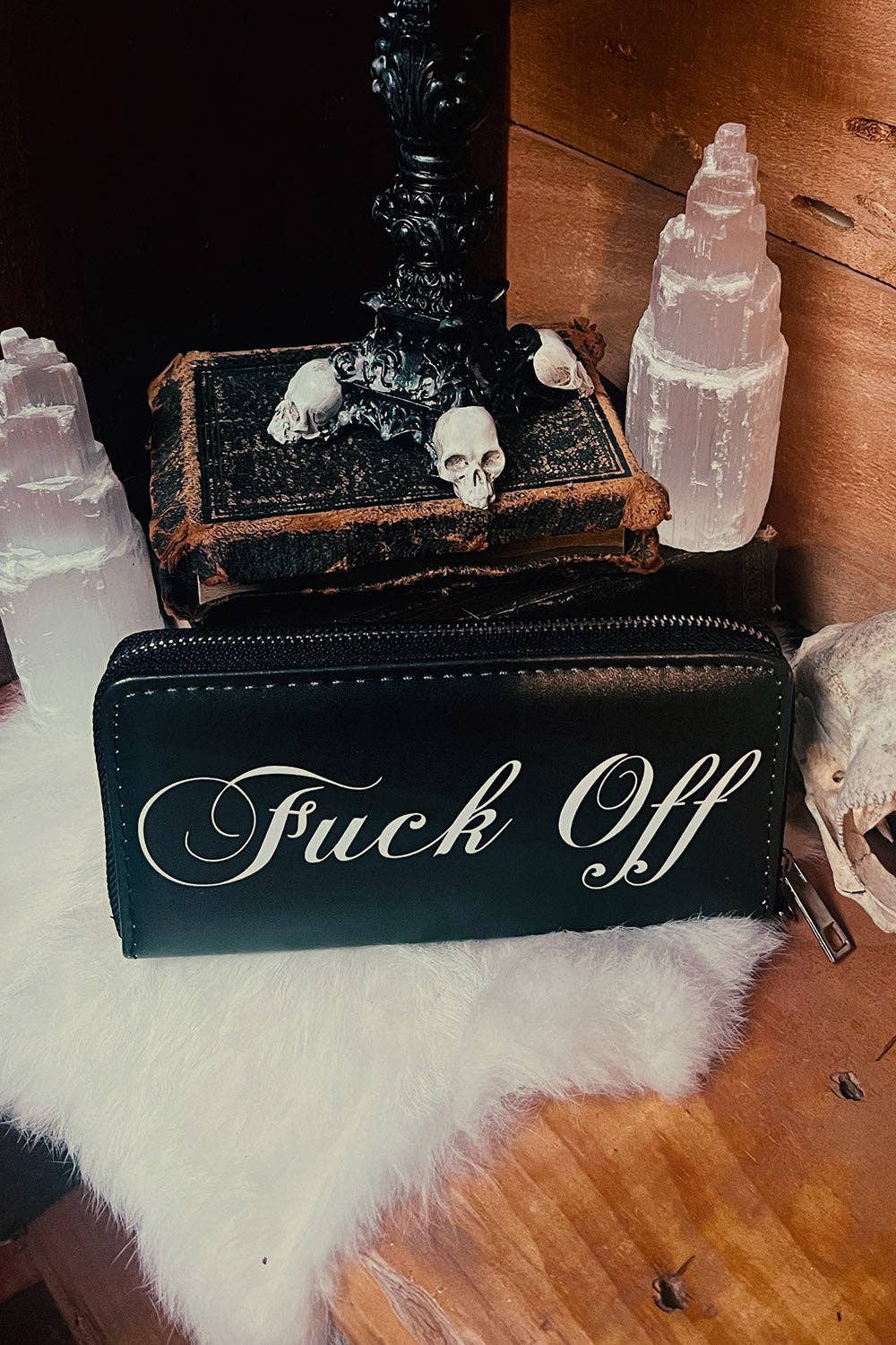 A black faux-leather zip pouch with the phrase 'Fuck Off' in white cursive script, displayed on a white faux-fur surface next to a black book with skull decorations and crystal salt lamps.