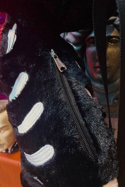Close-up of a black fuzzy bag  with a silver zipper and white curved stripe details, photographed against a dark background with blurred decorative objects.