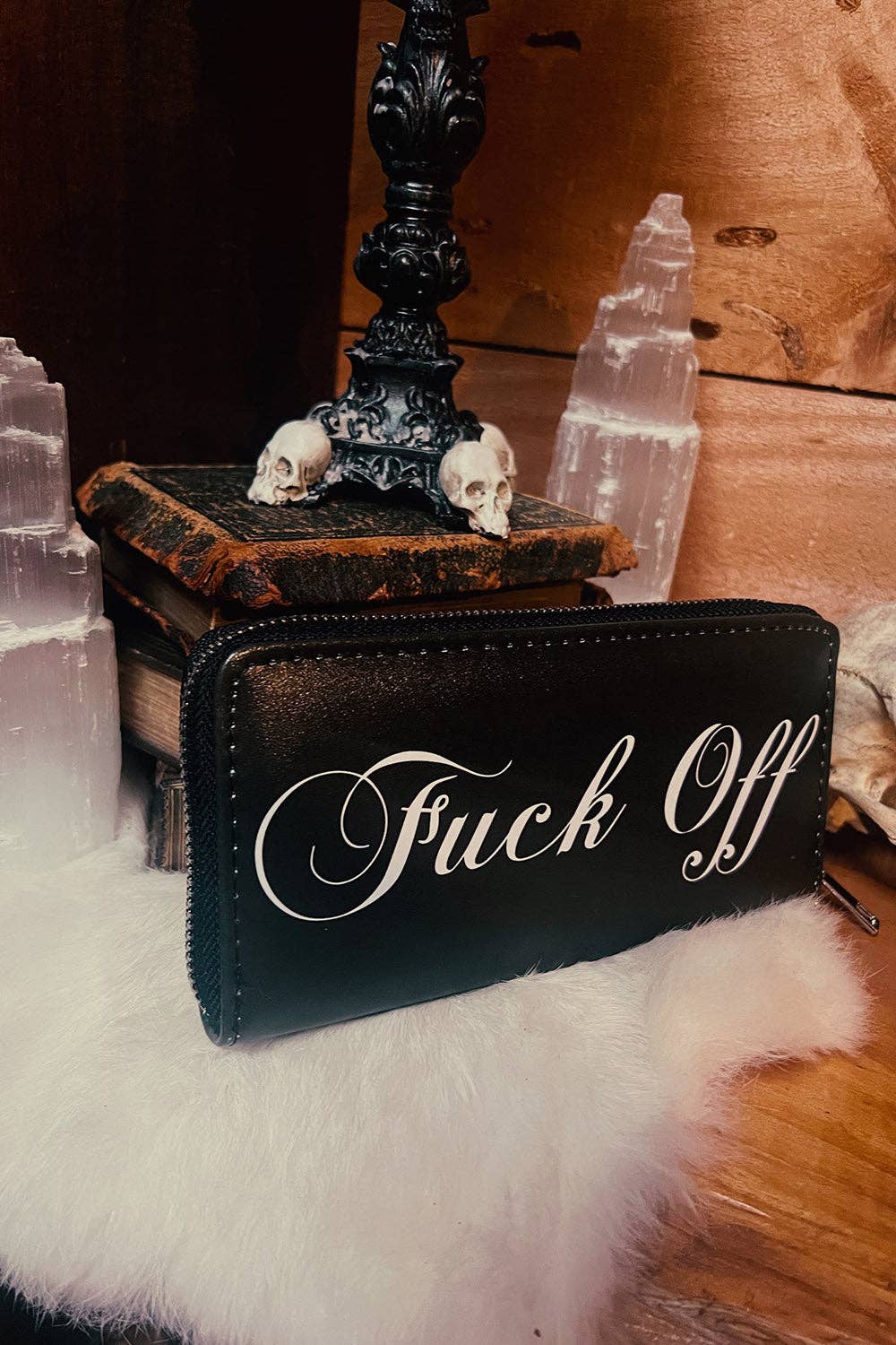 A black leather zip-around wallet with white cursive text reading 'Fuck Off' resting on a white faux-fur surface, surrounded by gothic props including skulls, old books, a black candlestick, and clear crystal towers against a wooden background.
