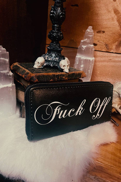 A black leather zip-around wallet with white cursive text reading 'Fuck Off' resting on a white faux-fur surface, surrounded by gothic props including skulls, old books, a black candlestick, and clear crystal towers against a wooden background.