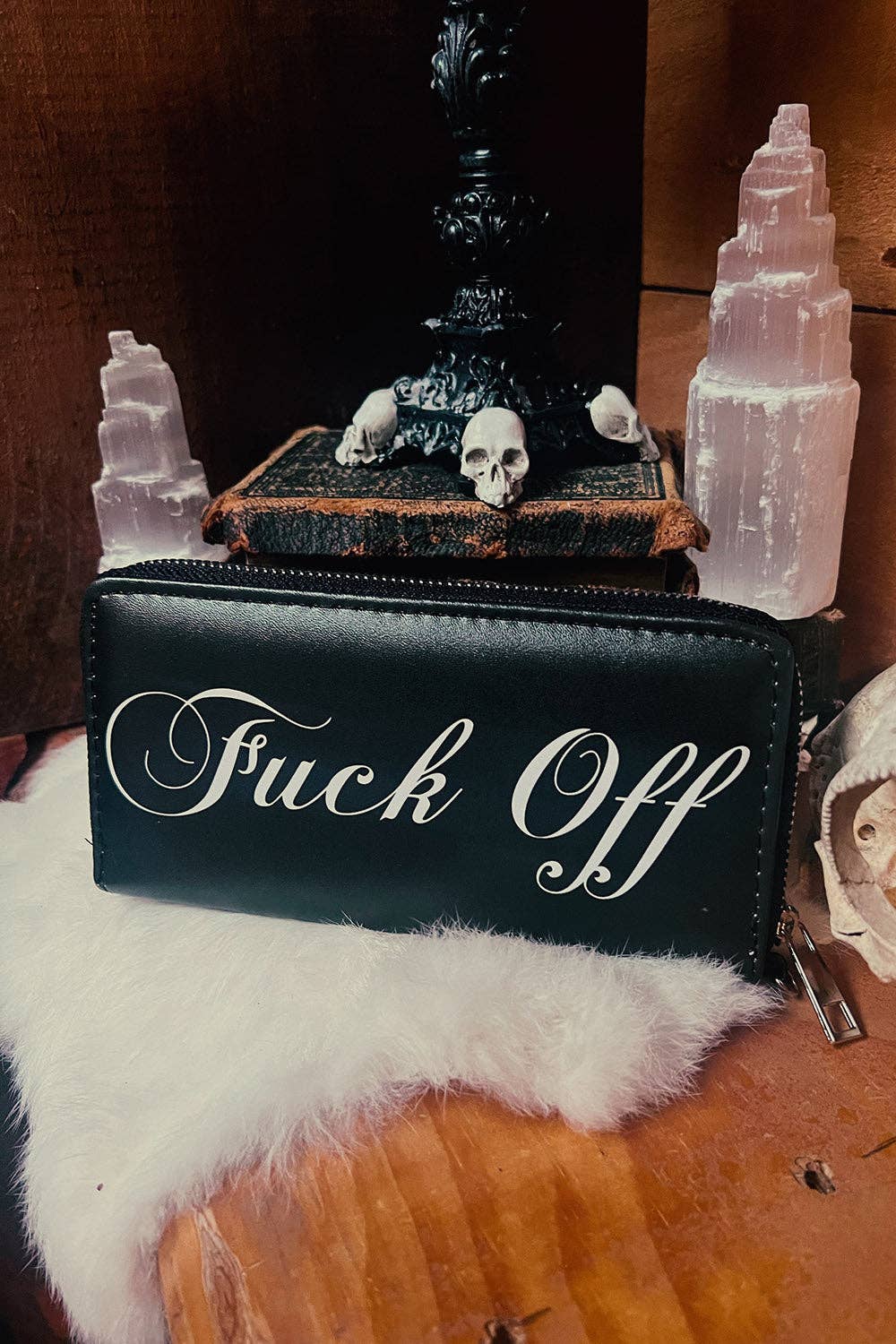 A black faux-leather zip-around wallet with white cursive text reading 'Fuck Off' on the front, styled on a white faux-fur surface with occult-themed props including a skull, old book, black candle, and pink crystal towers.