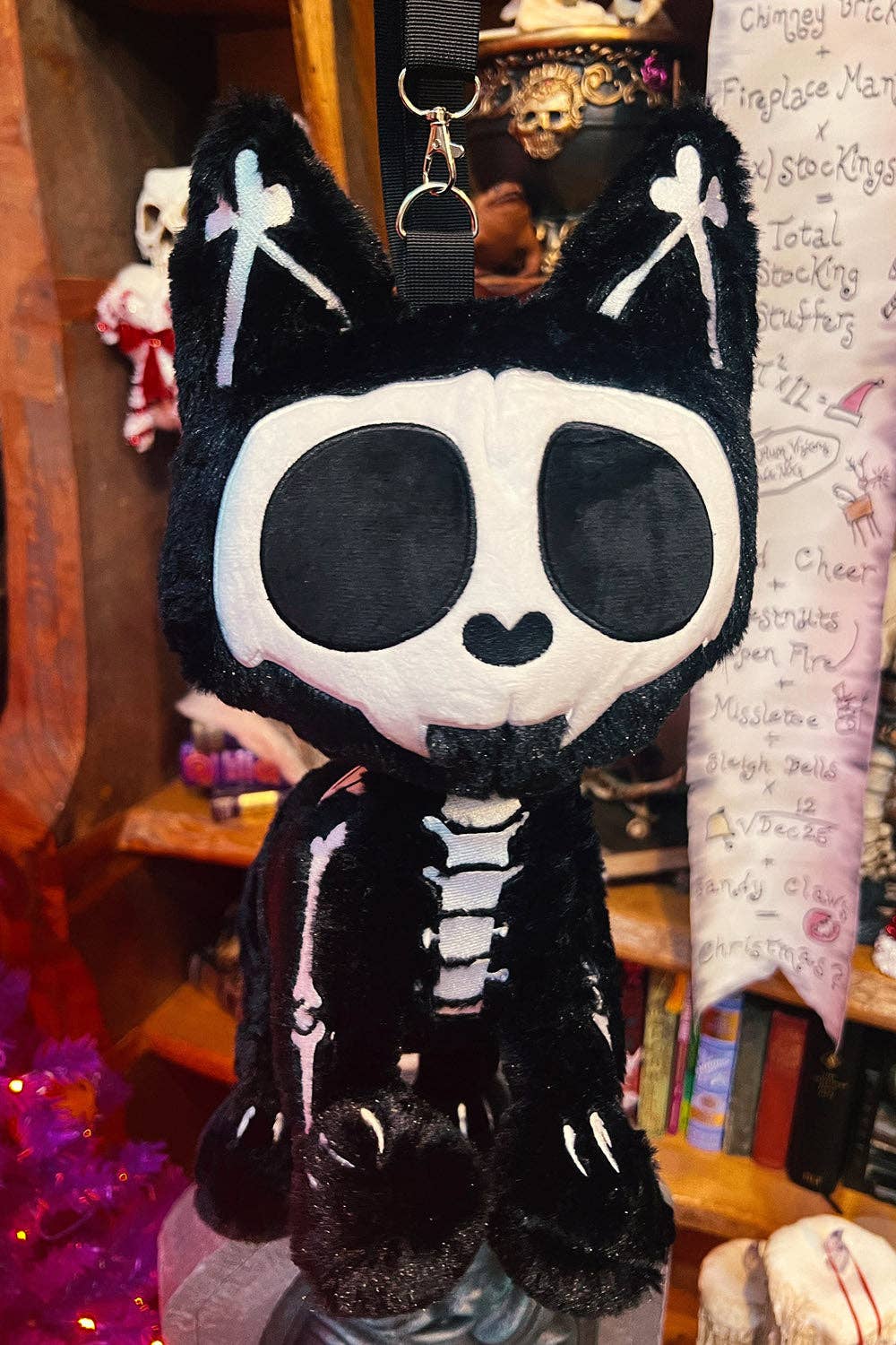 A black plush cat bag with a white skull face, white cross details on the ears, and a skeleton-print bodysuit, hanging in a shop setting.