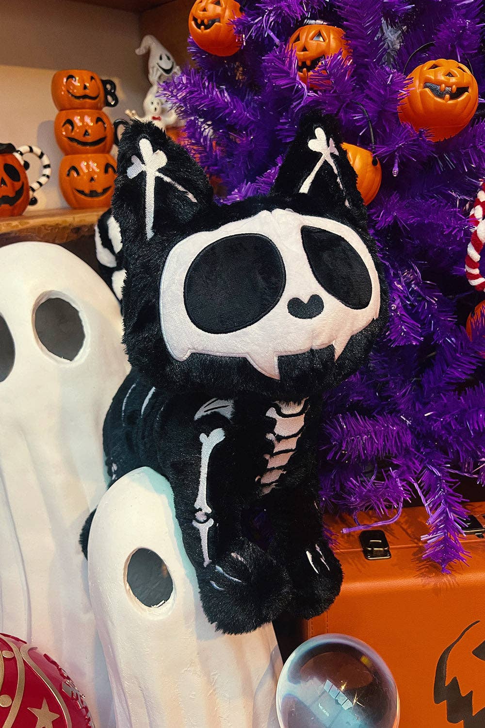 A black and white skeleton cat plush crossbody sitting on a white ghost-shaped pillow in front of a purple tree with pumpkin ornaments.