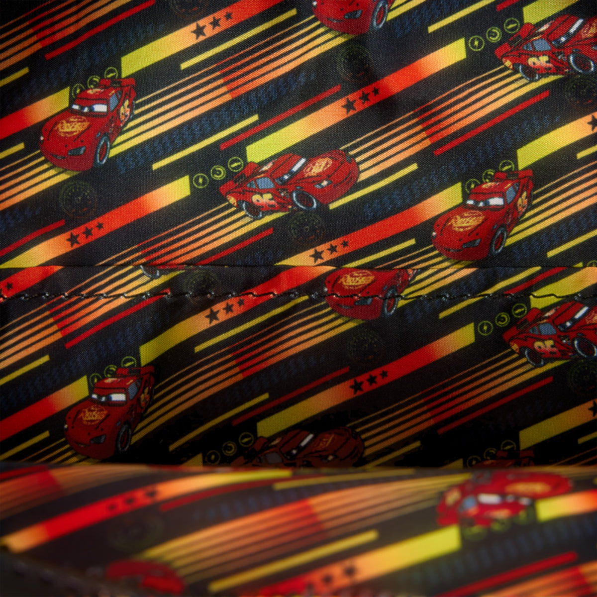 Close-up of a printed fabric featuring repeated red Lightning McQueen race car motifs over a black background with diagonal yellow and orange stripes.