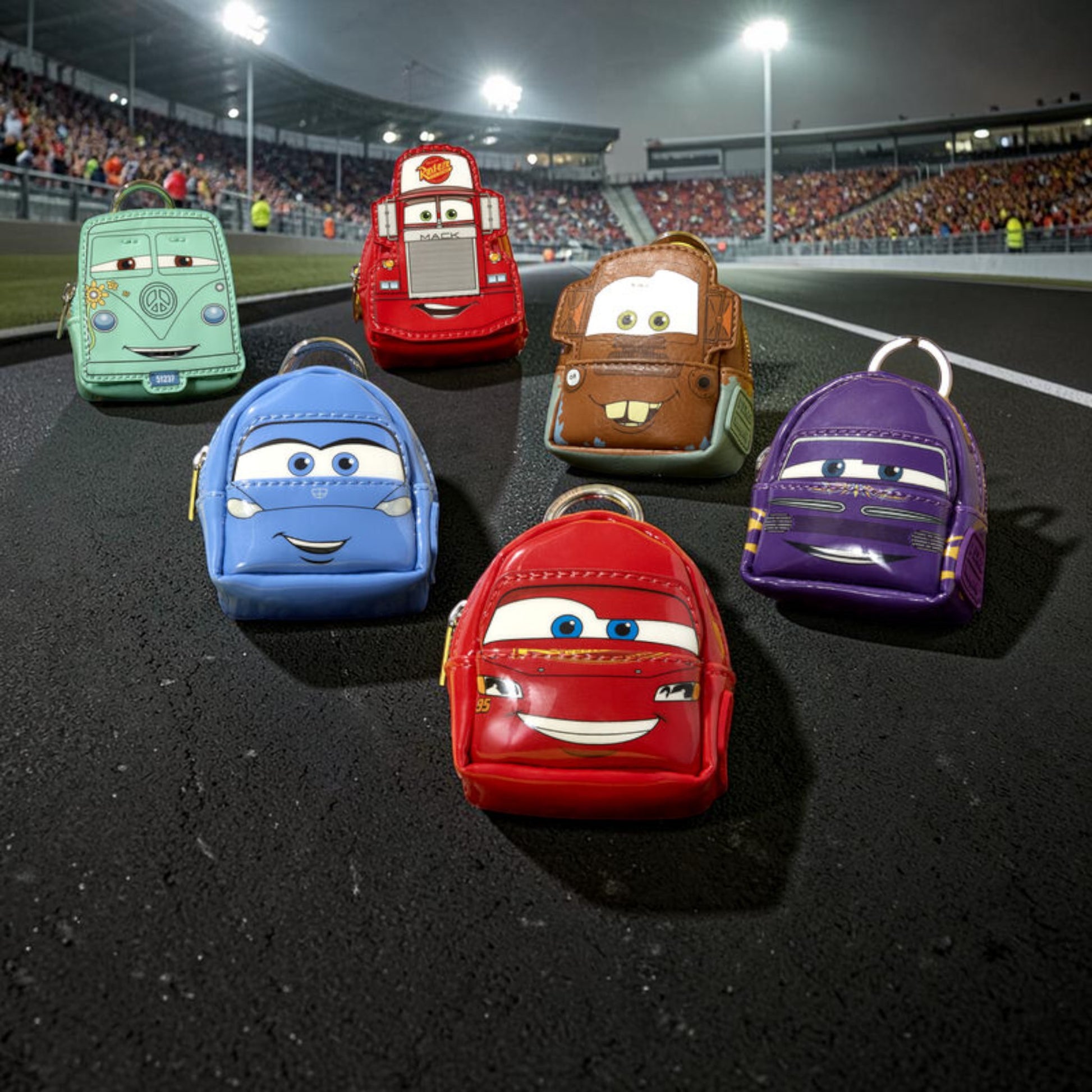 Five small character-shaped backpacks or pouches shaped like cartoon vehicles, arranged on a wet racetrack with stadium lights and a crowd in the background. The colors visible are mint green, red, blue, brown, and purple.