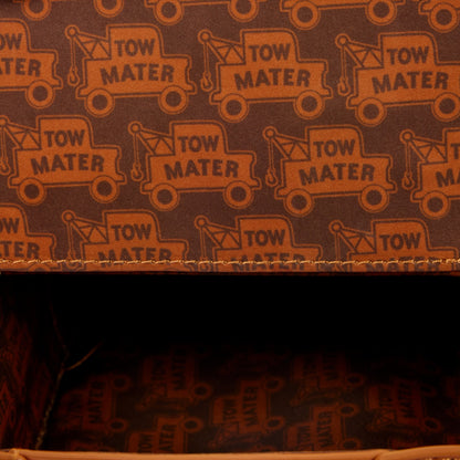 A close-up of a brown fabric or paper surface printed with a repeating orange tow truck pattern and the words 'TOW MATER'. The top edge shows a stitched seam, and the lower portion reveals a dark interior with the same printed material.