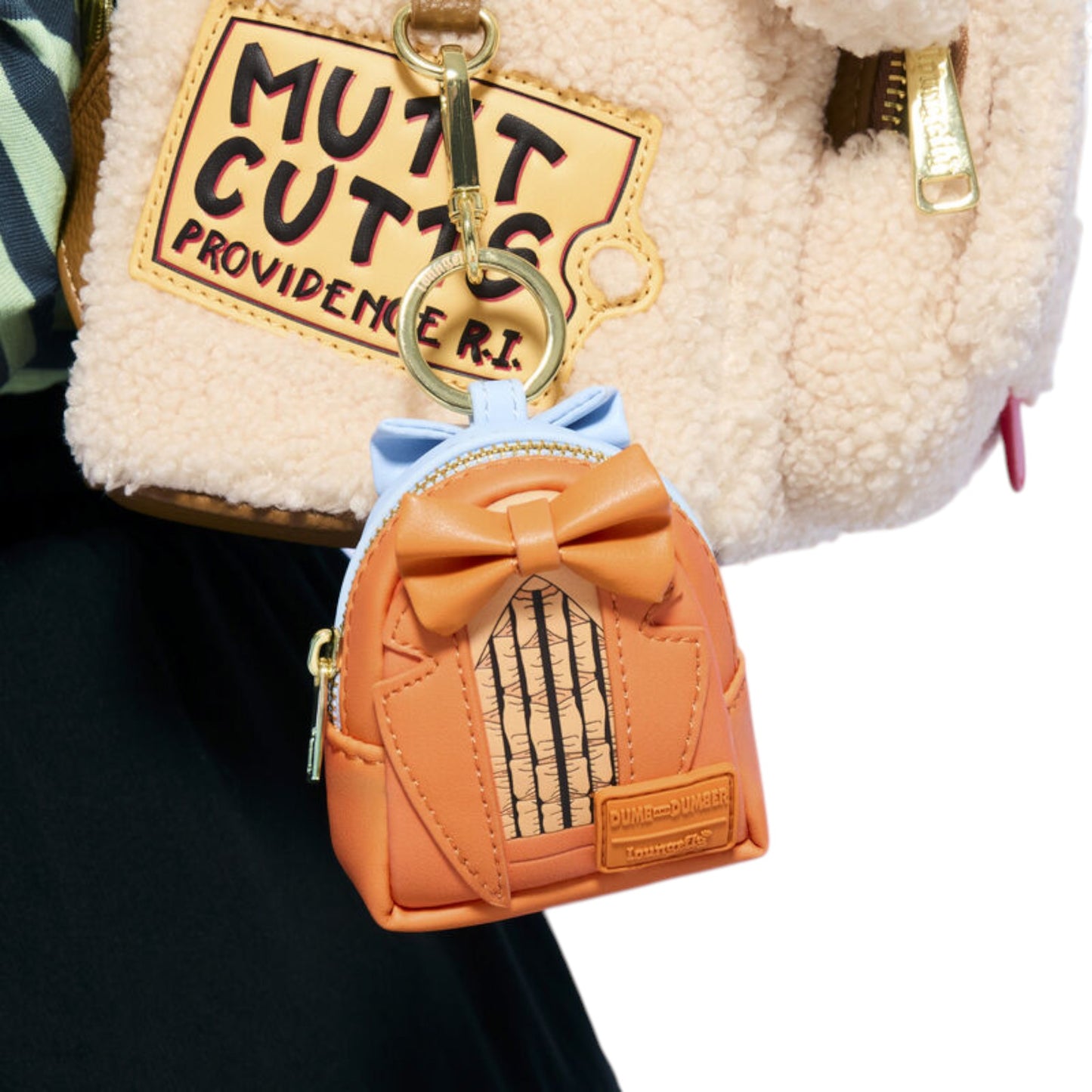 A small orange faux-leather mini backpack keychain charm with a bow and striped window detail, attached to a gold key ring and shown hanging from a cream sherpa bag.