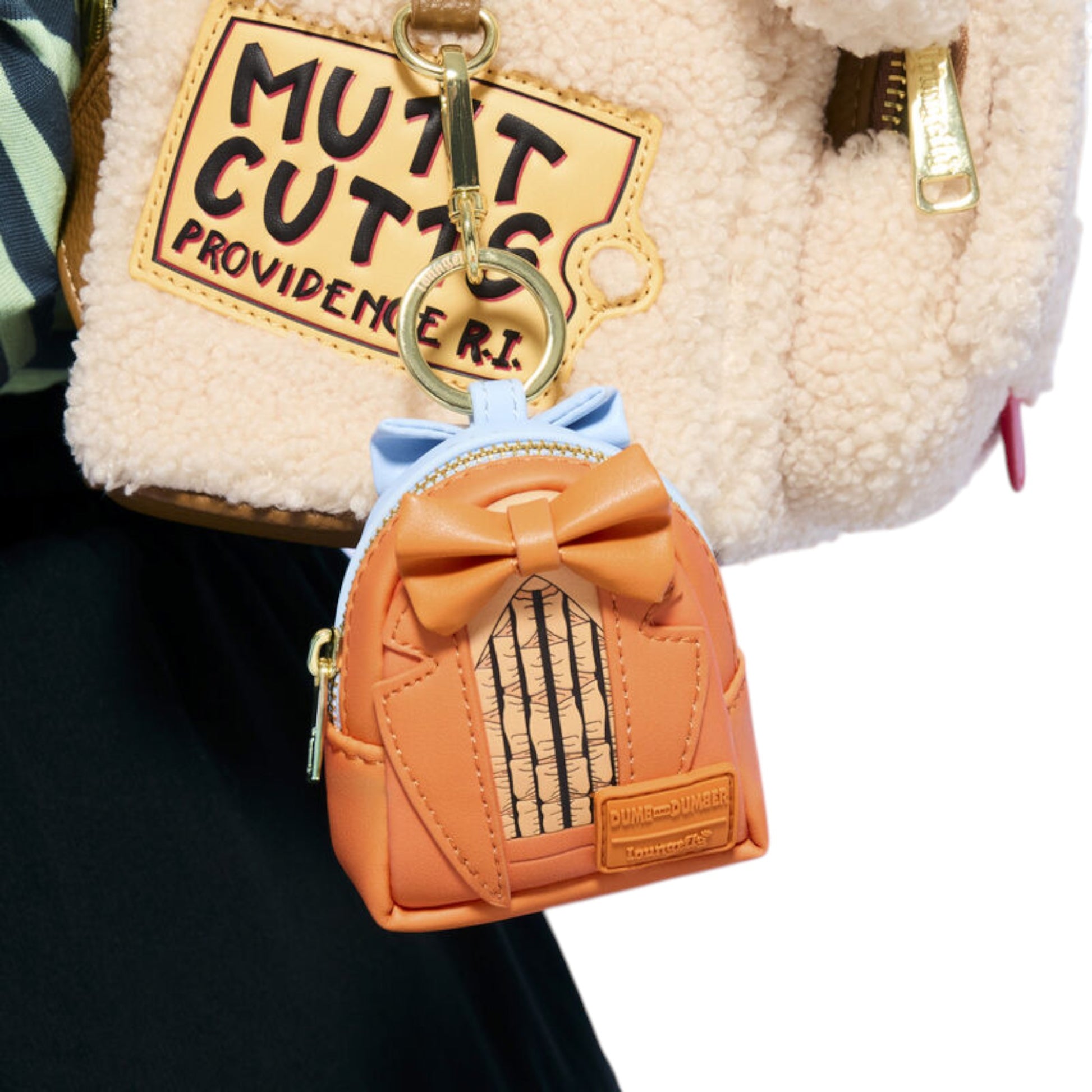 A small orange faux-leather mini backpack keychain charm with a bow and striped window detail, attached to a gold key ring and shown hanging from a cream sherpa bag.