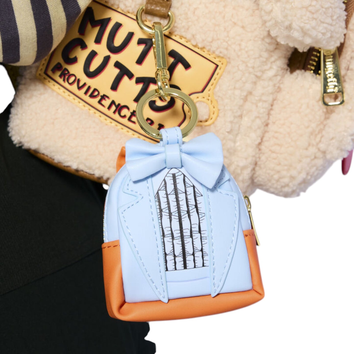 A small novelty keychain shaped like a tuxedo jacket with a light blue bow tie, light blue body, orange base, and black-and-white striped front panel, attached to a gold key ring and shown against a cream teddy-texture background.