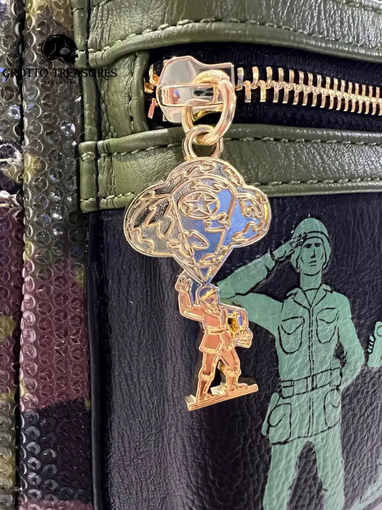 Close-up pf Camouflage Loungefly Disney Pixar Toy Story Army Men Sequin Mini Backpack with a gold and blue pendant zipper charm on a green textured surface with a soldier saluting design.