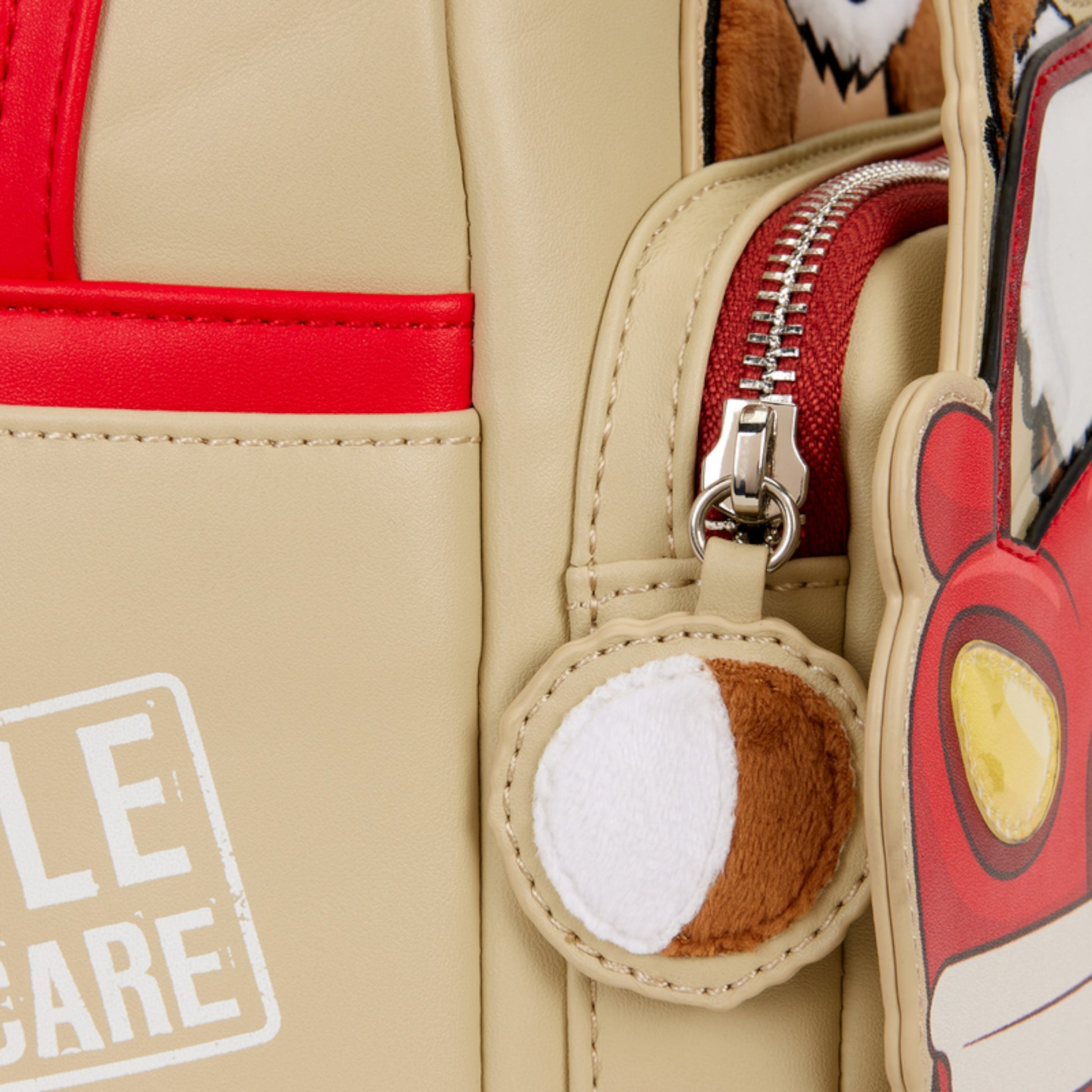 Close-up of a beige backpack with red accents, a gizmo character zipper charm, and a patch.