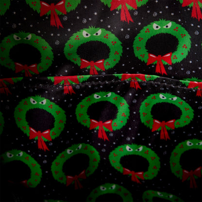 Close-up of a black fabric with a repeating pattern of green Christmas wreaths decorated with red bows and small red berries, with white snowflake details.