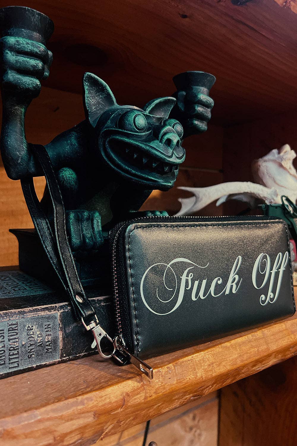 A black zip-around wallet or clutch with white script text reading 'Fuck Off' sits on a wooden shelf next to a dark green gargoyle figurine holding cups.
