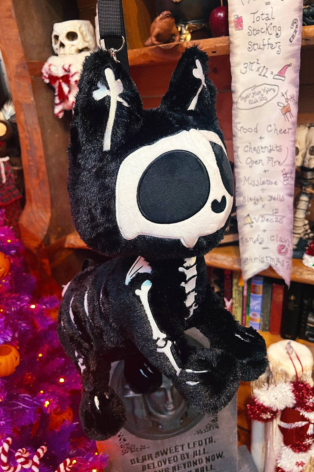 A black plush cat character bag with a white skeleton print and large black-and-white cat face, hanging in a decorated shop setting.