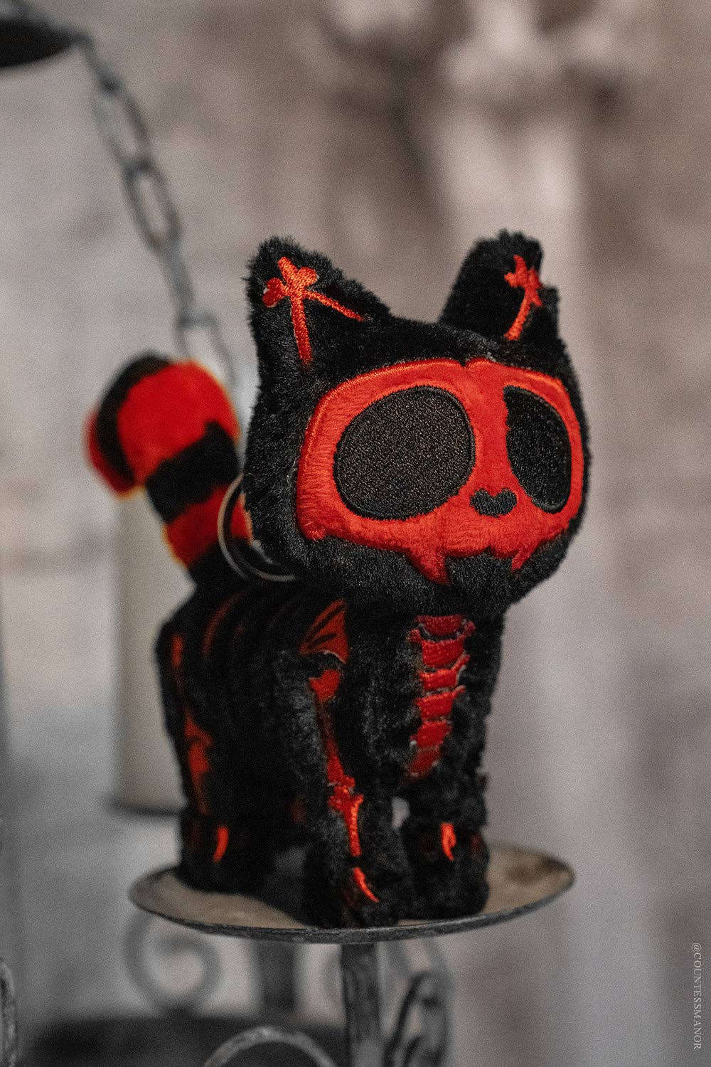 A black plush cat keychain with red embroidered skeleton details, skull face, and striped tail, displayed on a small metal stand against a blurred gray background.
