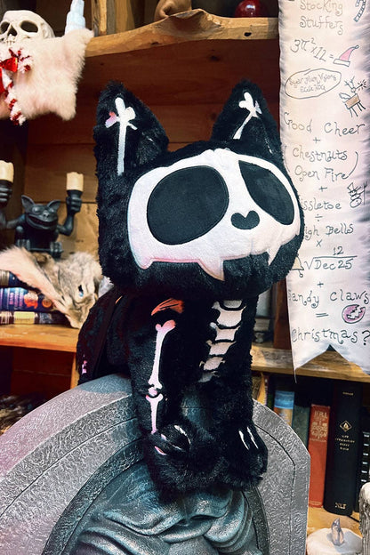 A black plush cat bag styled as a skeleton, sitting on a gray textured surface in a cluttered, cozy room with wooden shelves, books, and Halloween decorations in the background.