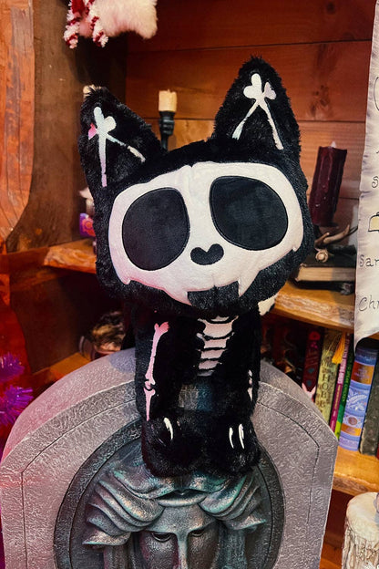 A black and white skeleton cat plush bag sitting on a gray decorative stone-like object, with a wooden shelf and books in the background.