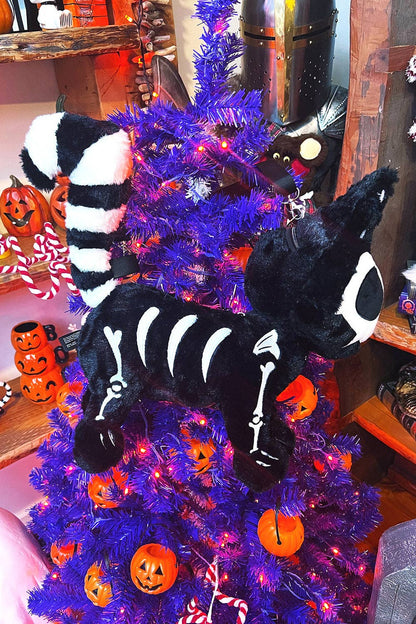 A black plush cat bag with white skeleton print and a striped tail, displayed on a purple Christmas tree with orange pumpkins and lights.