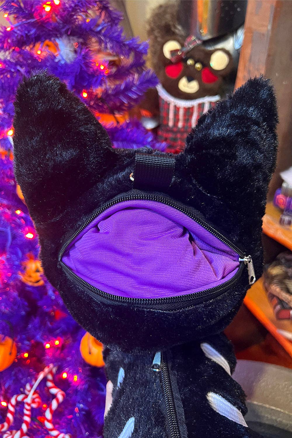 A black plush cat-shaped bag with pointed ears and a visible purple zippered interior, photographed close up against a festive purple holiday tree background.
